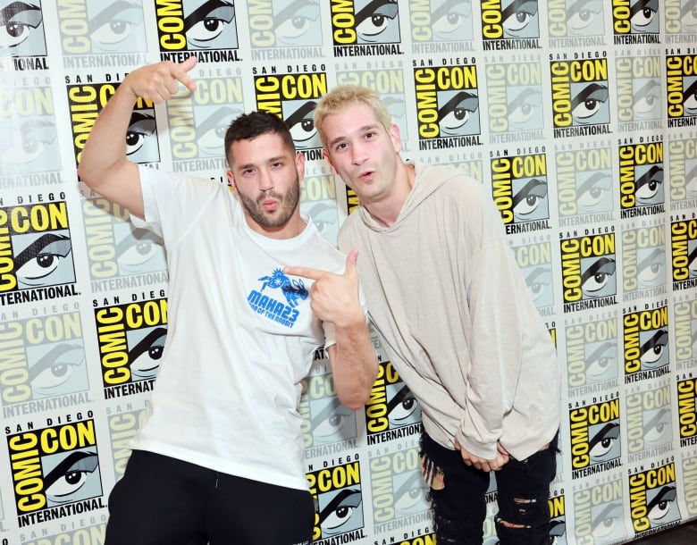 A man with dark hair and stubble gestures as he stands next to a man with blond hair in front of a wall decorated with San Diego Comic-Con logos.