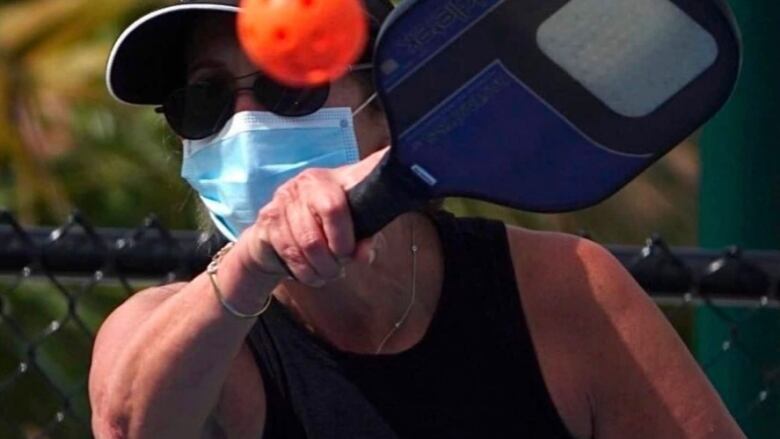 A woman wearing a ball cap, sunglasses and medical face mask swings a pickleball racket at a plastic wiffle ball.