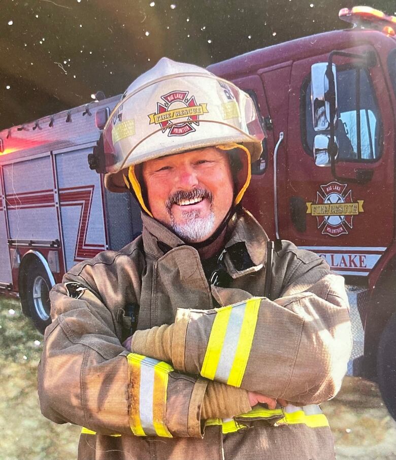 A man wearing firefighting gear stands with his arms crossed in front of a fire truck, smiling. 