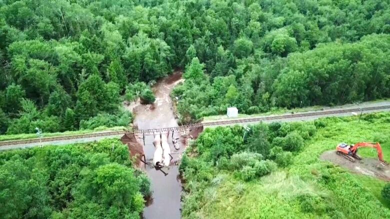 A damaged rail line is shown suspended over a river. There is forest on either side of the river.