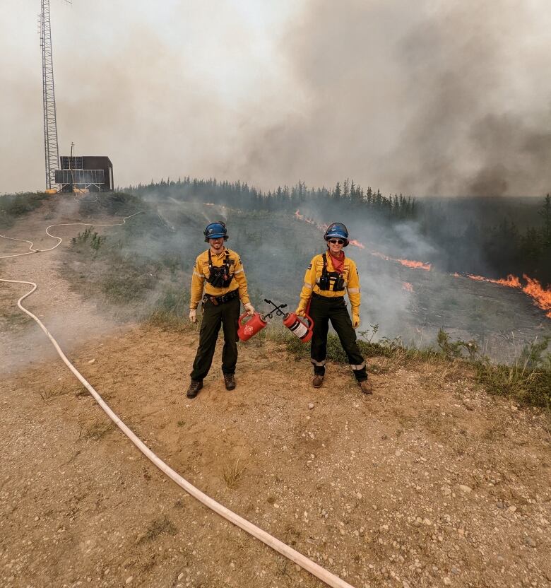 Two firefighters decked out in gear with a line of fire behind them.