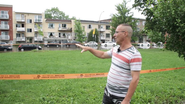 A man standing behind some orange tape gestures toward a partially-flooded street. 