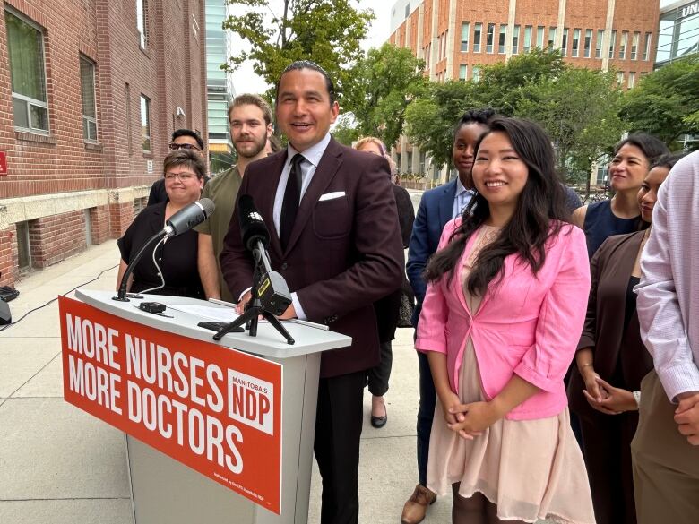 A man speaks behind a podium, while flanked by several fellow NDP MLAs, candidates and medical students.