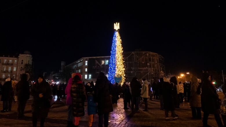 A night scene with people standing outdoors in front of a Christmas tree with the left side lit with blue lights and yellow lights on the right half. 