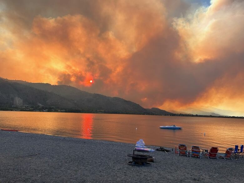 Plumes of orange smoke come from a hill over a scenic water body, with deck chairs and pool implements visible.