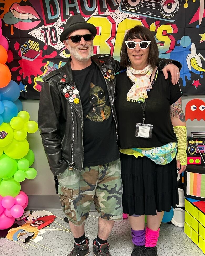 A man and a woman pose for a photo is 80s clothing with a brightly coloured poster in the background.