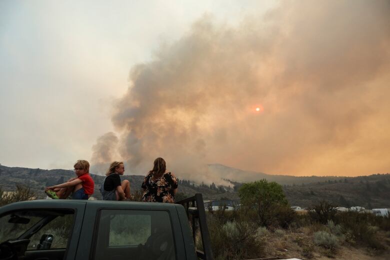 Three children sit on the roof of a dark green truck and watch a wildfire burning on a brown, desert hill in the distance. The sky overhead is orange as smoke obscures the sun.