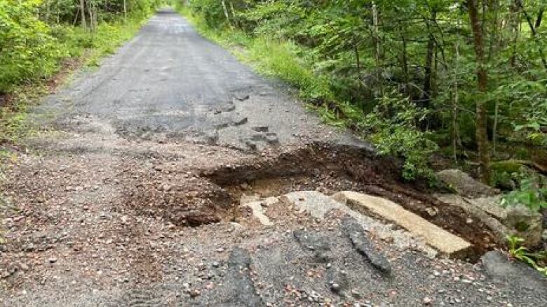 A cement pathway with a large pothole in its left side. There is gravel across the road and uneven rock within the pothole.