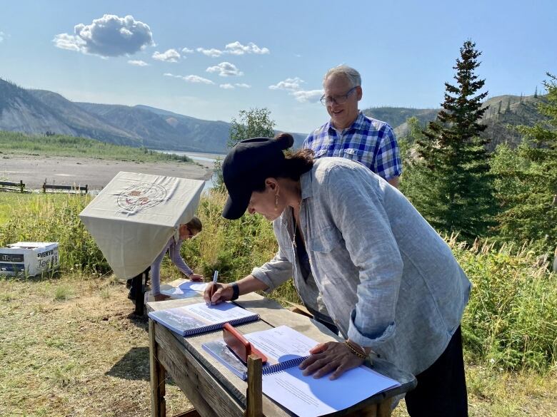 A woman stands outside a table signing a document as a smiling man looks on. Trees and mountains and a river are in the background.