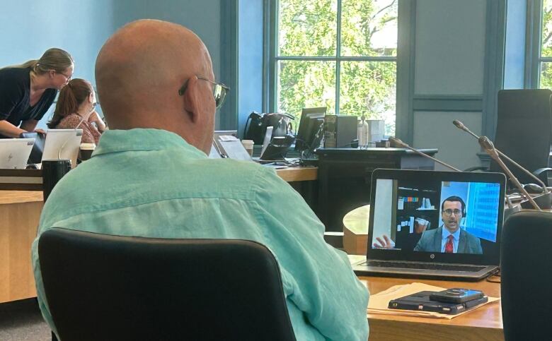 Bald man with glasses looks at computer where man in suit and tie is speaking