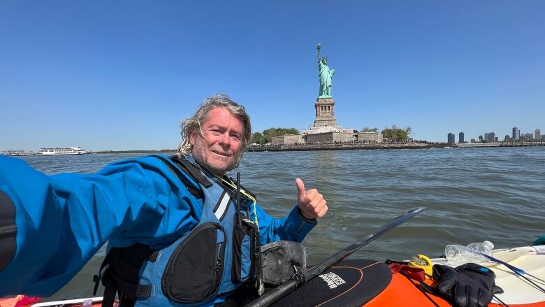 A man wearing a blue jacket sits in a kayak, smiles and gives a thumbs up. The Statue of Liberty in New York is seen in the background.