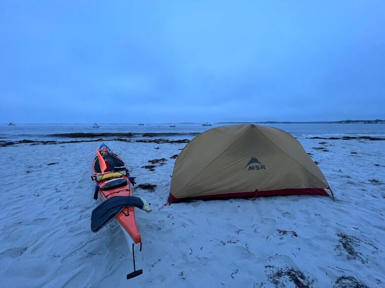 An orange kayak and a small tent sit on a secluded beach. The water is calm and the sky is cloudy.