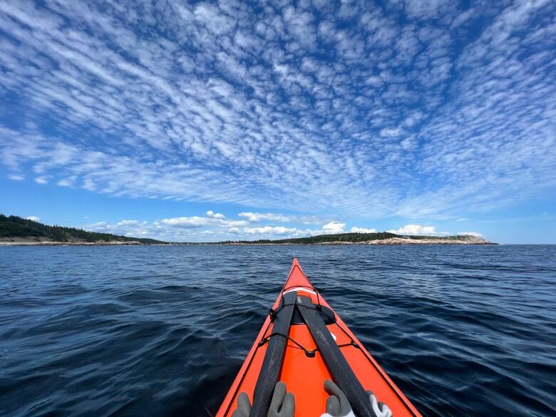 The tip of a bright orange kayak is seen on dark blue waters. The sky is blue but dotted with white clouds. Some beaches and green trees can be seen in the distance.