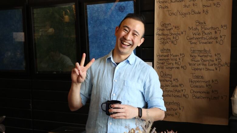 A man in a blue shirt holds a mug on one hand and makes a peace sign with the other.