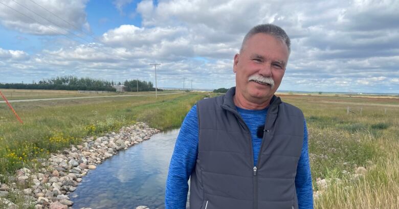 A man wearing a blue shirt and a black vest stands in front of an open-irrigation channel. 