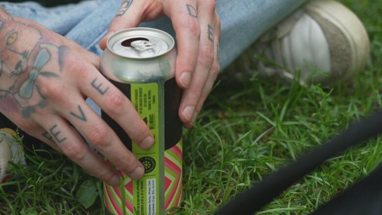 A can is shown held between two hands on grass.