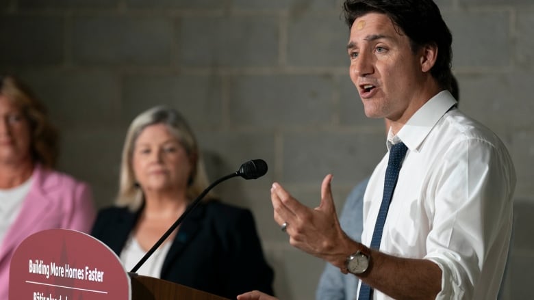 A man in a shirt and tie speaks at a podium while two women stand to the side.