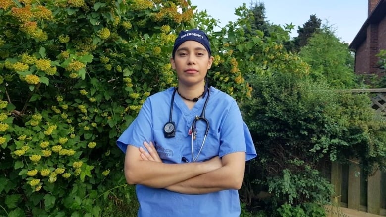 A nurse wearing blue scrubs, including a navy scrub cap and a stethoscope around her neck, stands with her arms crossed in front of some bushes.