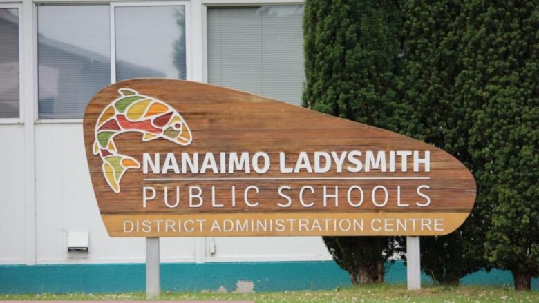 A crafted wooden sign featuring a small orca logo in a rainbow of colours that says Nanaimo Ladysmith Public Schools is seen on a lawn with a hedge to one side and school offices behind it.