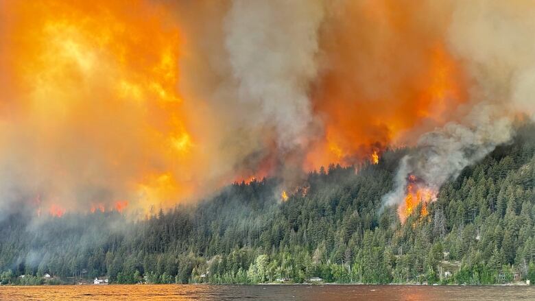 Plumes of smoke and licks of flame are seen on a hillside next to a picturesque lake.