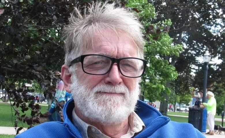 A man with a white beard, white hear, black sunglasses and a blue sweater. 