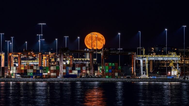 A huge orange moon sits above a port loading dock illuminating shipping containers and reflecting in the black ocean water.