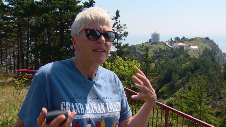 A woman with short, gray hair, wearing sunglasses stands with expressive hands gestured out in front of her. Behind her is a large, green peninsula covered with trees. In the distance, there's water. 