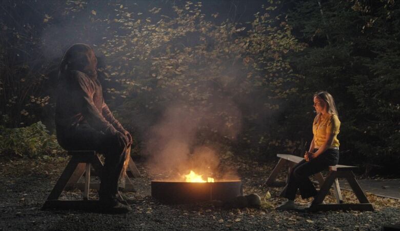 A still from a film shows a hooded man and a young woman sitting facing each other on opposite sides of a campfire.