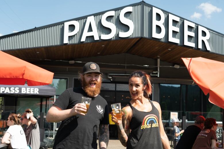 A man and woman in black shirts stand smiling holding glasses of beer