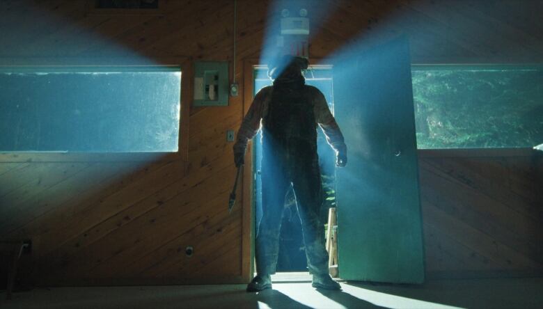 A still from a film shows a large, backlit man wearing a hood and holding an axe standing in the doorway of a cabin.