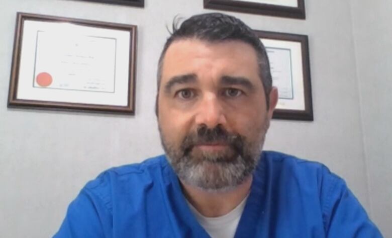 A veterinarian wearing blue scrubs sits at his desk with diplomas hanging on the wall behind him.