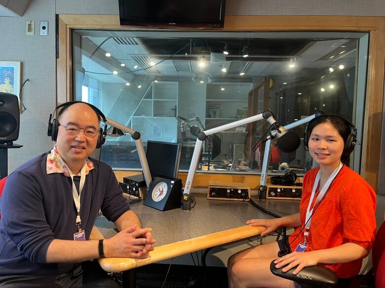 A man and a woman sitting at an expansive desk, both wearing headphones, pose for the camera in a radio studio with large glass windows and microphones on boom cables.