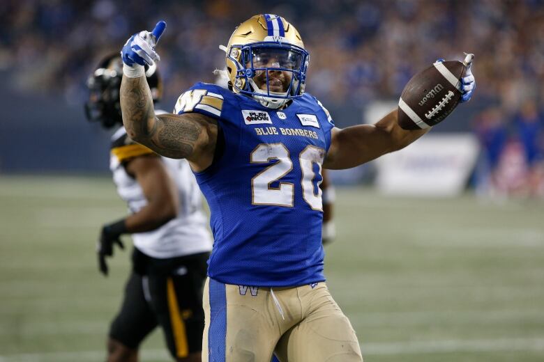 A football player in a blue jersey smile and raises his arms in celebration while holding a football in his left hand.