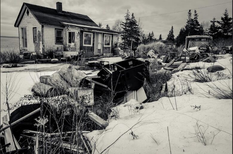 A black-and-white photo of an old house in winter. There's wood and debris strewn about the yard.