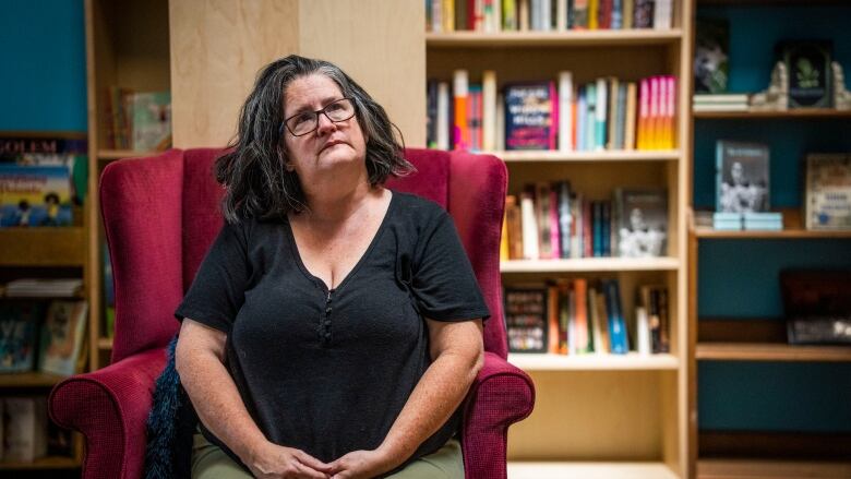 A middle aged woman sits in a red armchair with book shelves behind her. She wears green pants and a grey short-sleeved shirt. Her hair is shoulder-length and a mix of grey and black and she wears black glasses.