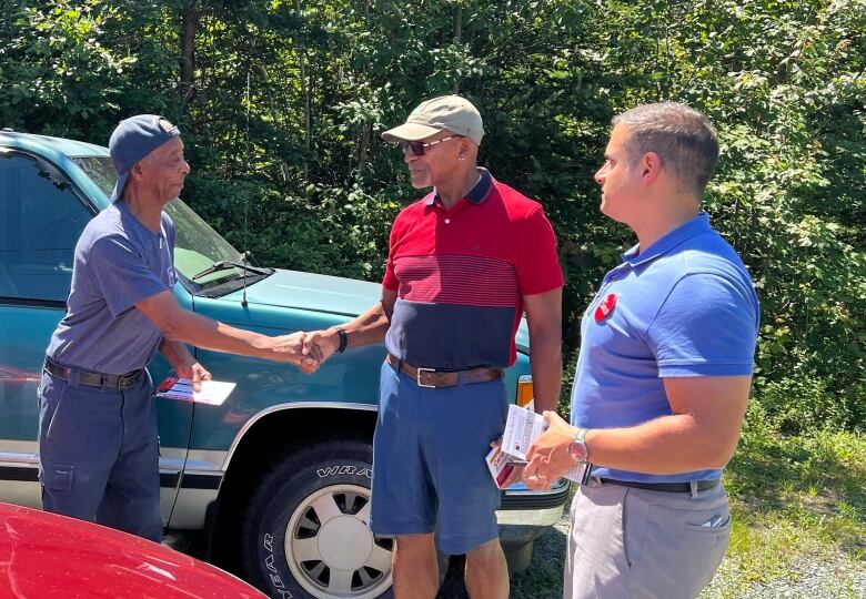 Three men are shown, two of them shaking hands, in between a green vehicle and a red vehicle.