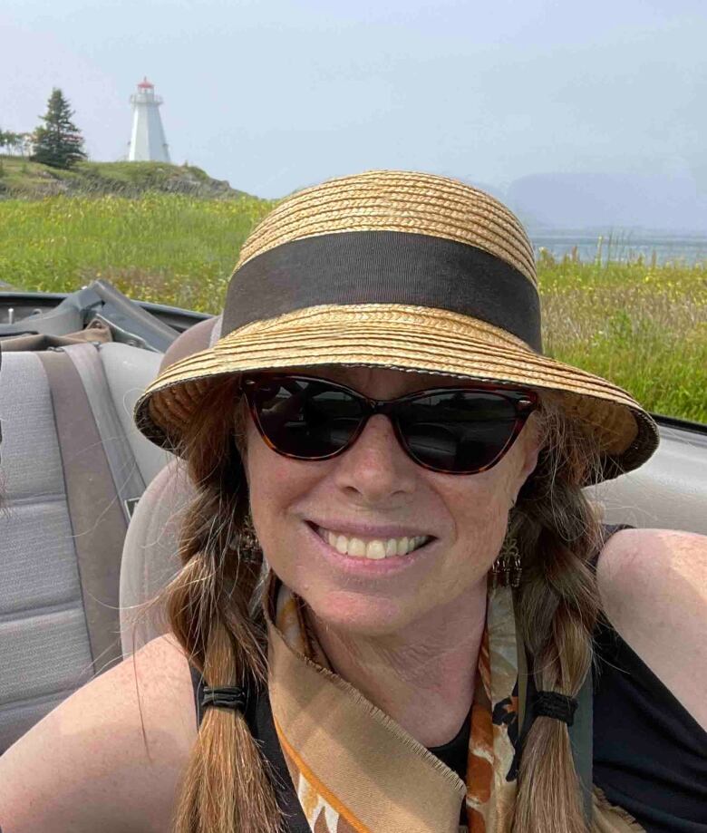 A woman with braided hair, sunglasses and a sun hat sitting in a car with a lighthouse in the background.