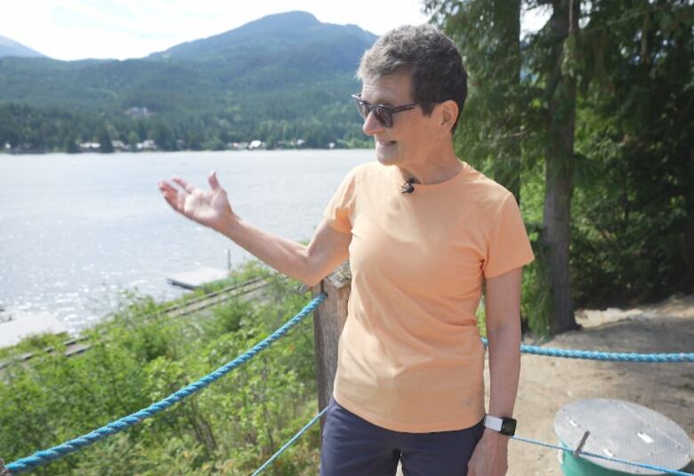 A middle-aged woman wearing blue shorts, an orange shirt and black sunglasses stands on a patio gesturing to the lake sparkling in the sunlight below her.