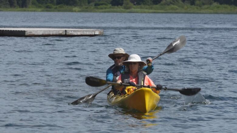 A yellow kayak paddled by a woman in the front and a man in the back seat glides across a lake. Both paddlers where grey tilley hats, black sunglasses and black lifejackets.