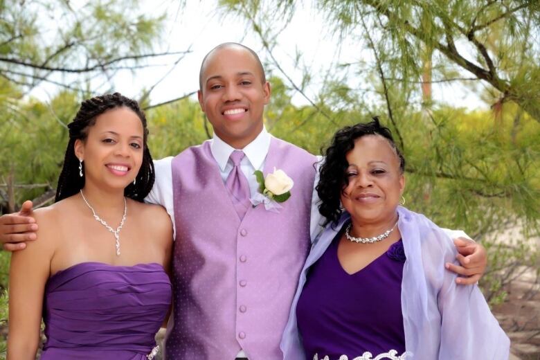 Three people are smiling wearing formal, purple clothing. Two people are wearing necklaces and one is wearing a tie.