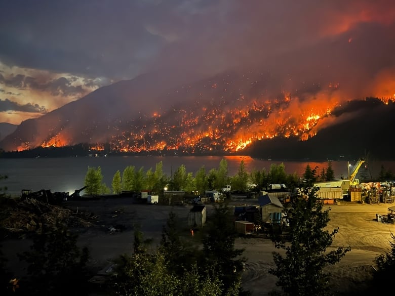 The side of a hill at the edge of a lake is consumed by flames casting an angry, red glow on the night sky.