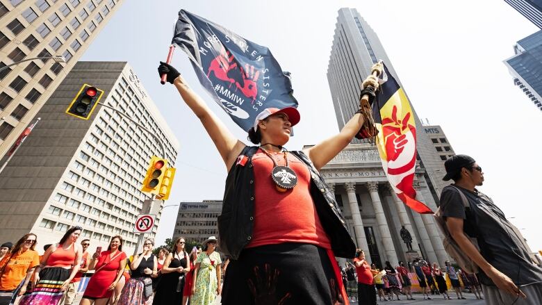 A woman holds up flags at the middle of Portage and Main, while other people prepare for a round dance.