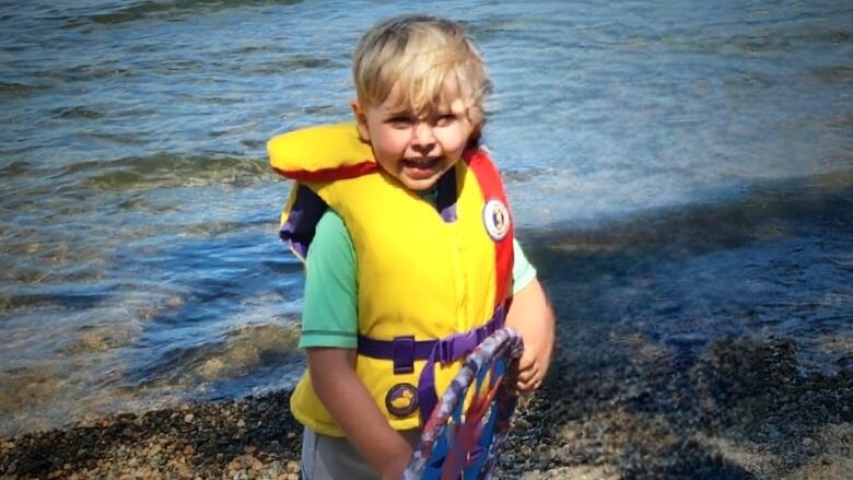 A boy with blond hair and a yellow life jacket stands at the water's edge.