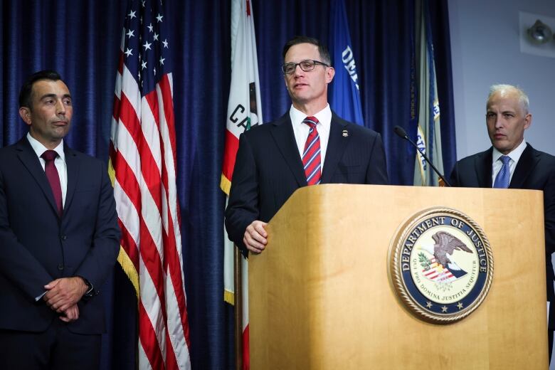 A man in a suit is shown at a podium speaking, with two other men in suits looking on in indoors in a room with curtains and a series of flags include the U.S. flag in the background.