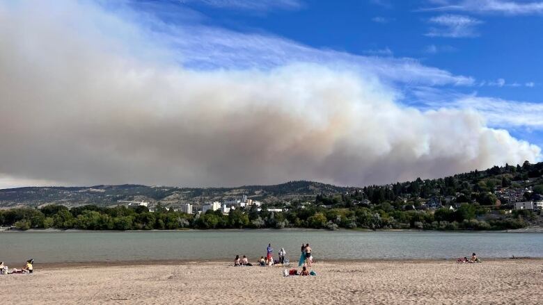 Wildfire smoke is seen rising from a hillside on the far side of a lake on a summer's day. A group of beachgoers is pictured on the sand in the foreground.