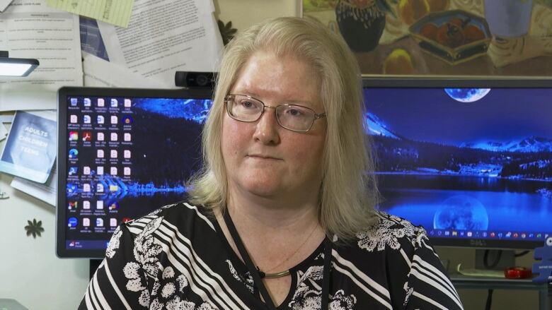 A woman with shoulder-length blond hair and glasses in front of two computer screens with nighttime photos as the screensavers. 