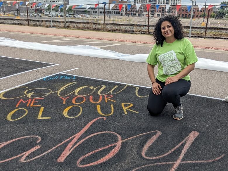 A woman with shoulder-length dark, curly hair kneels on the street, on a black painted section of her calligraphy painting on the street surface. She's wearing a lime-green tee-shirt, black pants and sneakers.