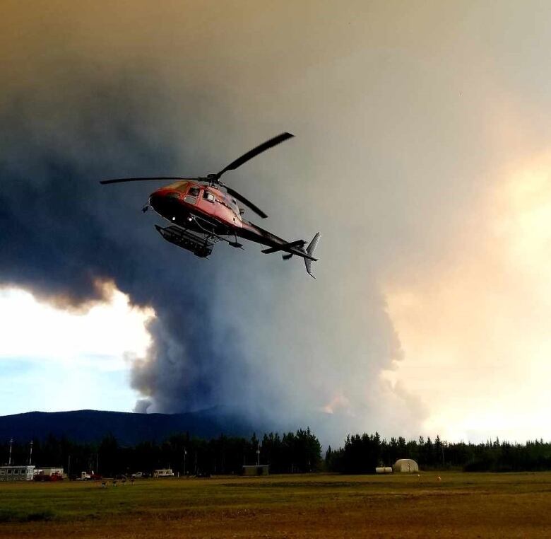 A helicopter is seen taking off with a big plume of wildfire smoke visible in the background.