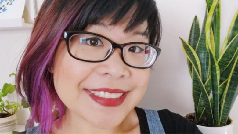 A smiling Asian woman with black-rimmed glasses and pink highlights in her hair with some plants in the background