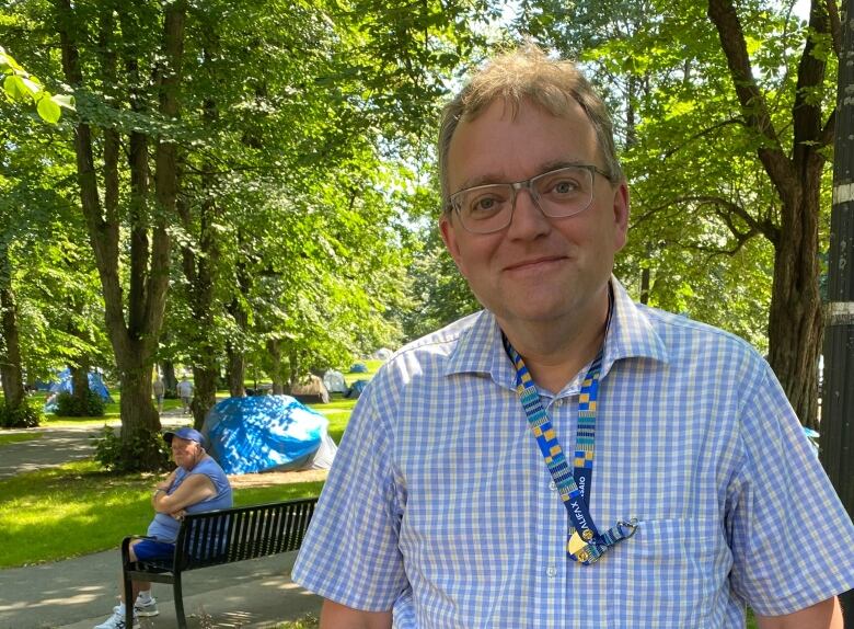 A mab wearing glasses and a blue shirt stands in a park and smiles slightly at the camera.